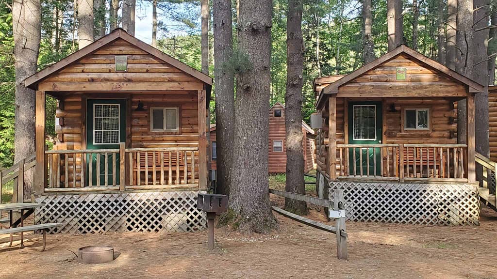 Two log cabins surrounded by blooming flowers in a wooded area perfect for a Spring getaway.