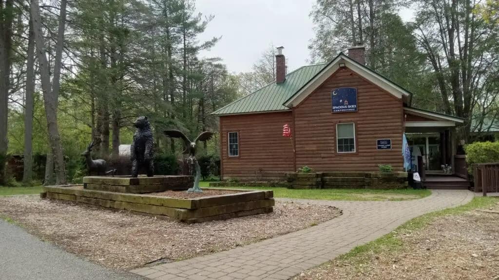 A wooden building with a green roof is surrounded by trees and wildflowers. A bear statue, another animal statue, and a winged statue are situated next to the building on brick pathways.