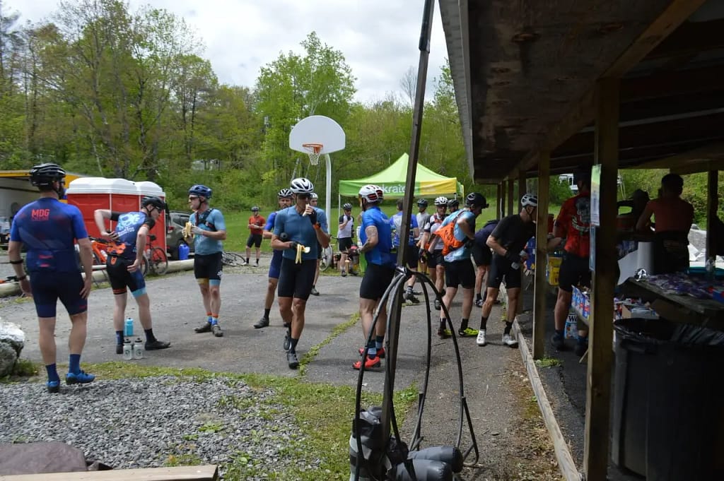 Several cyclists in sportswear gather near a building with outdoor equipment, portable toilets, and a basketball hoop during a break in a bike event amidst the scenic backdrop of Woodland Hills.