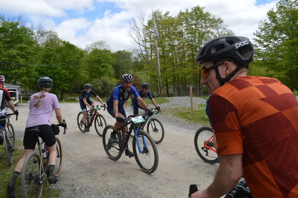 A group of cyclists on a gravel path, wearing helmets and biking gear, ride through a park area with trees in the background, embracing the scenic beauty of a Nature Lover's Journey.