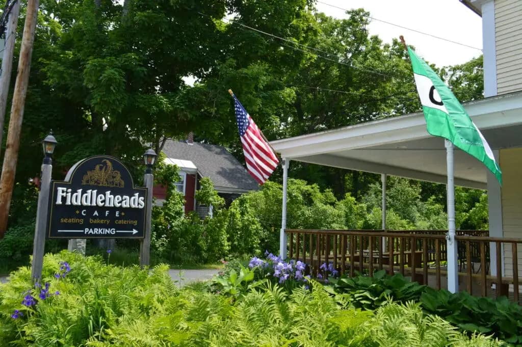 Exterior view of Fiddleheads Café in Hancock NH with an American flag and a green flag on the porch, surrounded by lush greenery and purple flowers. A sign for parking, perfect for those visiting from Spacious Skies Seven Maples Campground, is also visible.