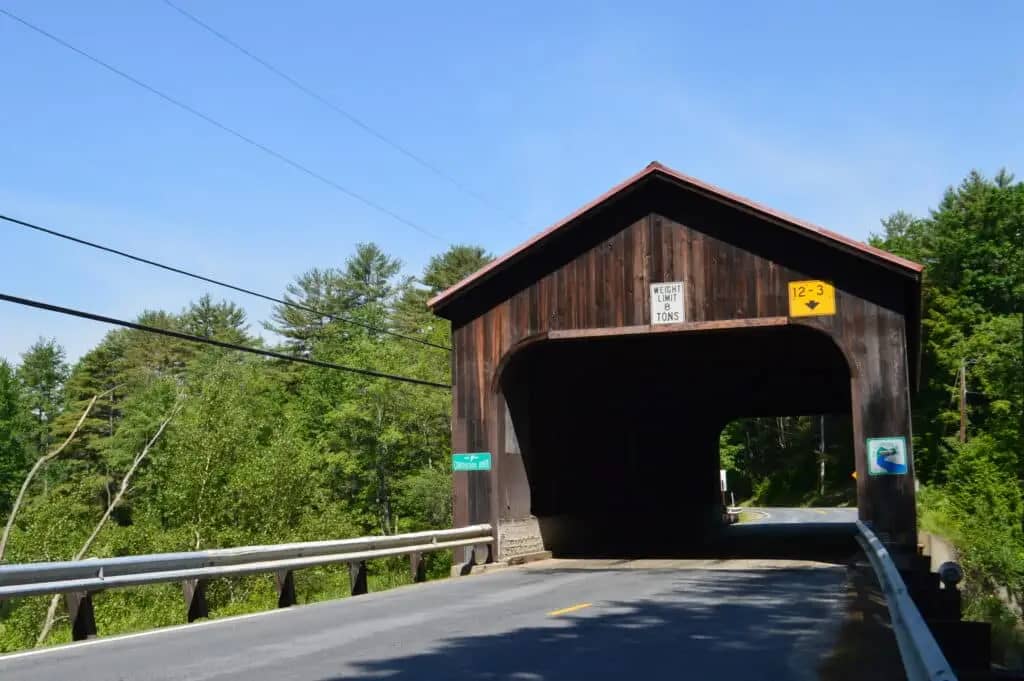 A wooden covered bridge with road signs indicating height and weight limits stands near the entrance to Spacious Skies Seven Maples Campground in Hancock, NH. The road passes through the bridge, surrounded by green trees under a clear blue sky.