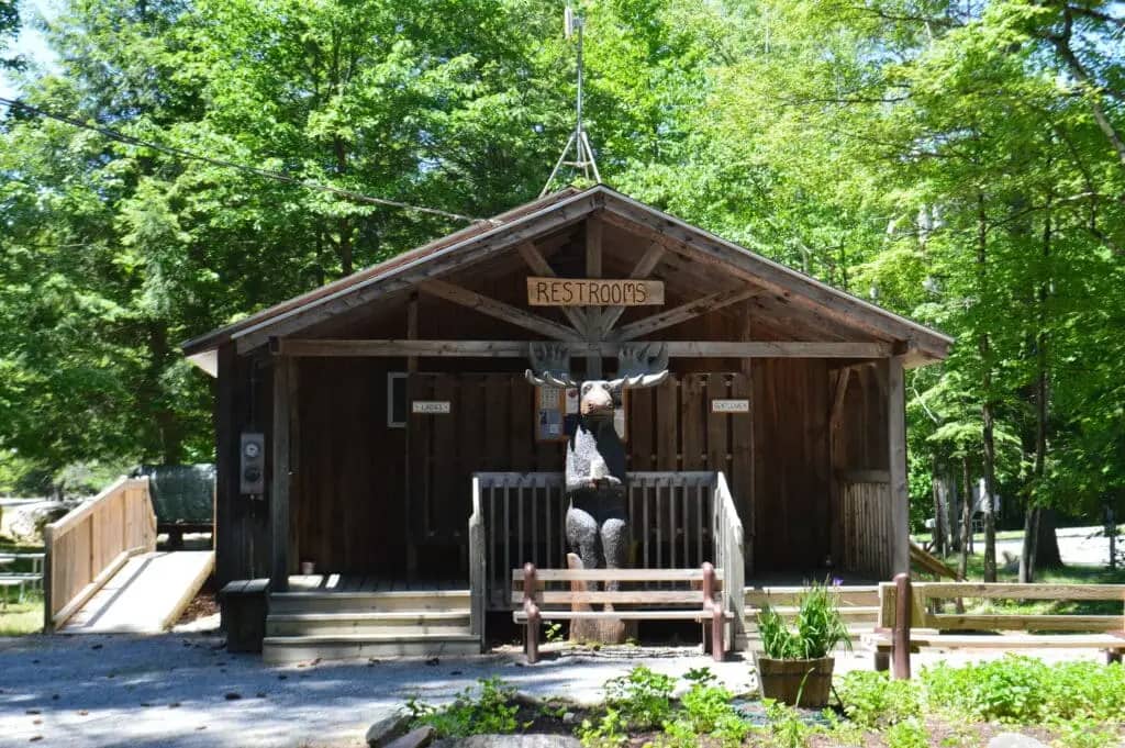 A wooden restroom building with signs indicating restrooms and a moose statue in front, nestled among trees along a gravel path, evokes the rustic charm of Seven Maples Campground in Hancock, NH.