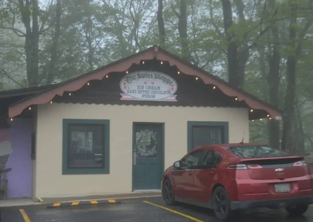 A small shop with a sign reading "The Swiss Shoppe - Ice Cream, Hand Dipped Chocolates, Fudge." A red car is parked outside. The shop is surrounded by trees and wildflowers, creating a picturesque scene in the wooded area.
