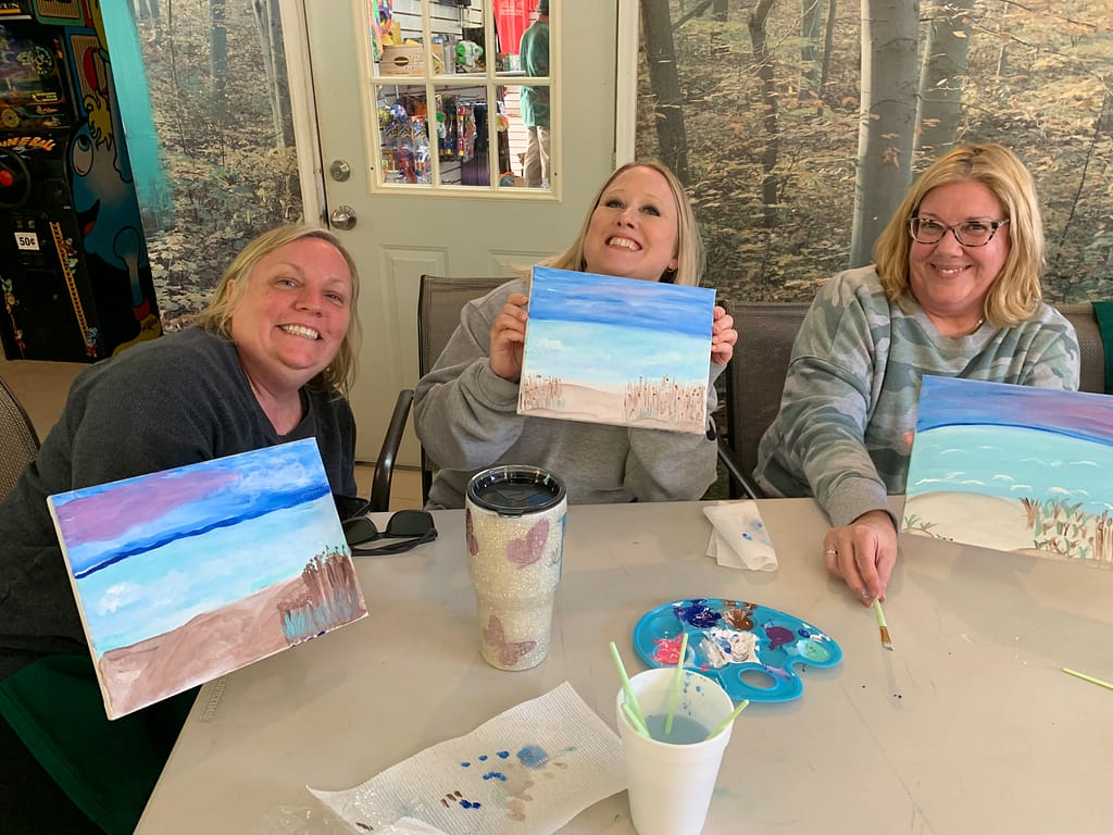 Three women smiling and holding paintings with beach scenes at a table, ready for their Springtime Adventure. The table is filled with art supplies, including paint, brushes, and cups. In the background, posters and a door complete this creative scene.