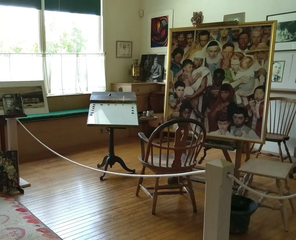 An art studio in New York State with a large painting of diverse people, an easel, wooden chairs, and various artworks displayed. Sunlight filters through windows with green blinds, casting a serene glow. A rope barrier separates the area. It's a perfect stage for Rustic Revelations.