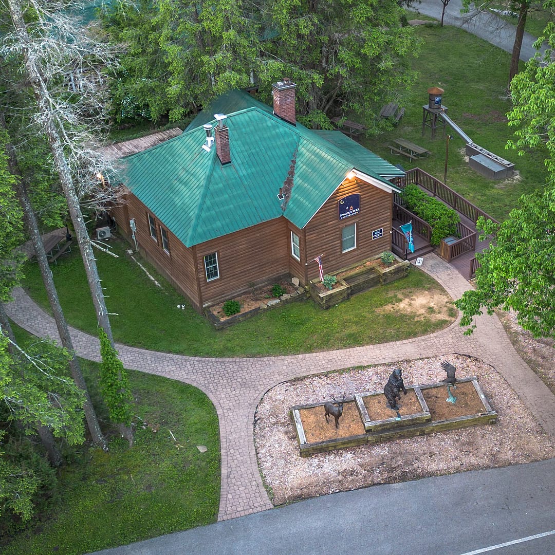 An aerial view of Belle Ridge cabin with a green metal roof, surrounded by trees and a paved path. A small playground is to the right. There is a memorial with three dark-colored statues on the lawn near the entrance, and American flags are displayed outside the cabin.