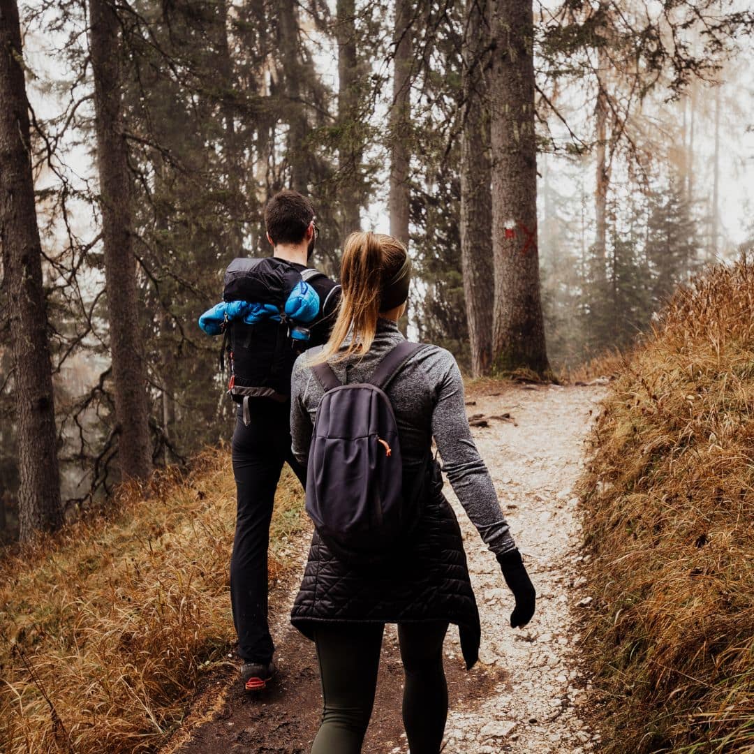 Two people hiking on a forest trail surrounded by tall trees and fog, feeling the love of nature embracing them. One wears a backpack with a blue jacket attached, the other carries a dark backpack. The path beneath their feet is lined with dried grass and leaves, as if guiding them through hidden galaxies of tranquility.