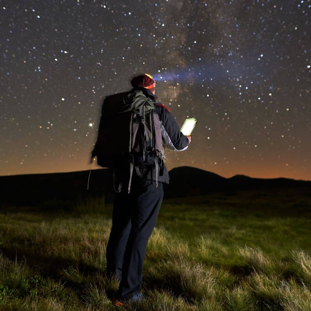 A person stands in a grassy field at night, holding a flashlight and looking up at a clear, star-filled sky. They wear a backpack and outdoor gear, casting a silhouette against the horizon—a moment of wonder reminiscent of finding Love Among the Galaxies.