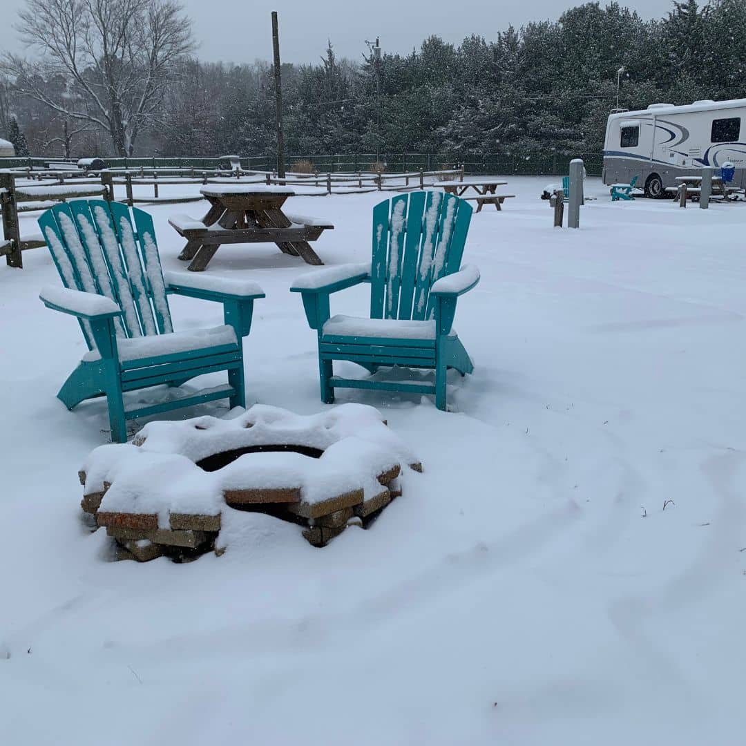 Two turquoise Adirondack chairs surround a snow-covered fire pit in a snowy landscape. In the background, a picnic table and camper trailer are set against snow-dusted trees. The scene is serene and wintry, evoking love for nature's quiet beauty amid galaxies of snowflakes.