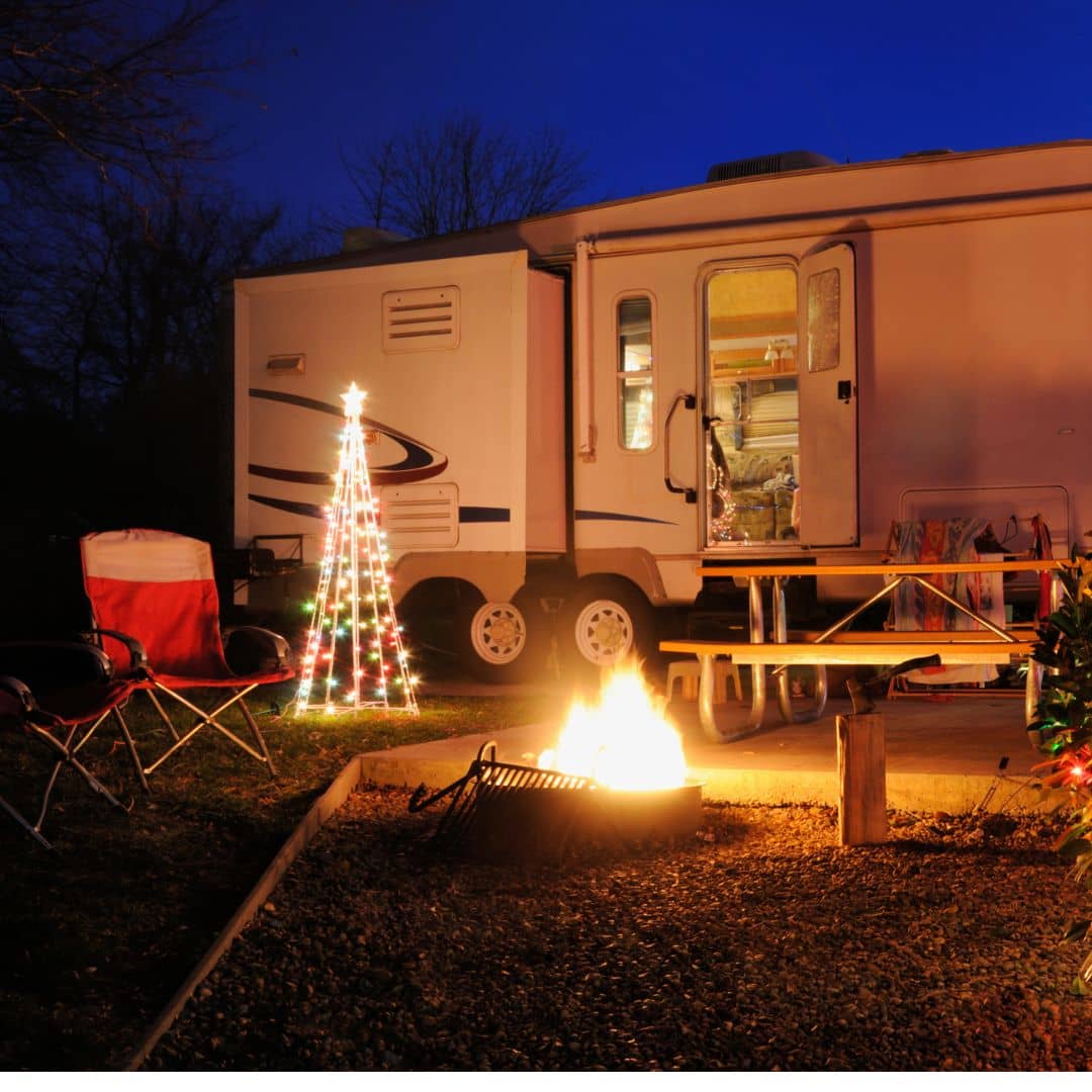 A cozy campsite scene with a trailer illuminated from within. "Love Among the Galaxies" is symbolized by a Christmas tree decoration, glowing near a crackling campfire and folding chairs. The evening sky is deep blue, creating a warm and inviting atmosphere.