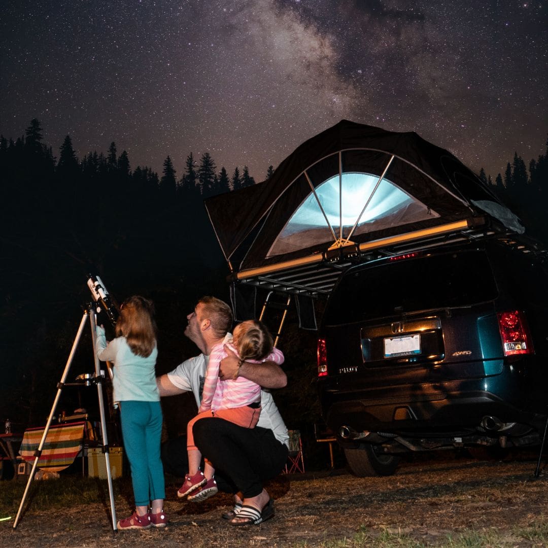 A family is camping under a starry night sky, wrapped in the warmth of love. A man kneels beside two children, one eagerly holding a telescope to explore distant galaxies. A car with a rooftop tent is parked nearby, and trees are silhouetted against the bright stars of the Milky Way.