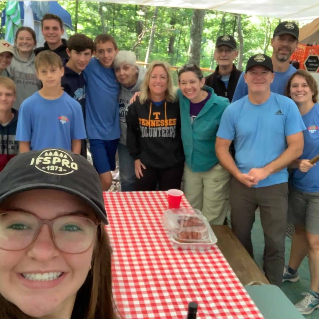 A group of smiling people gathered at a campsite with trees in the background. They stand around a table with a red and white checkered tablecloth, a dish of food, and camping gear visible. The scene is filled with love as they enjoy a casual outdoor event under the vast sky filled with galaxies.