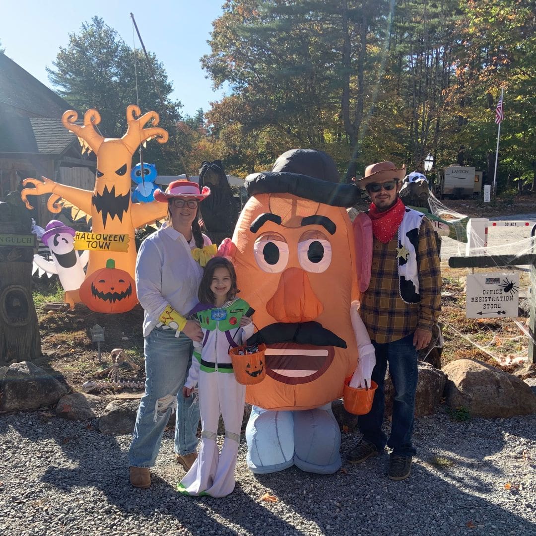 A group of three people in Halloween costumes stand outdoors. A child dressed as Buzz Lightyear holds a pumpkin bucket, ready to collect candy. They are surrounded by inflatable Halloween decorations, including a Jack-o'-lantern and a large Mr. Potato Head, as if celebrating love across galaxies.