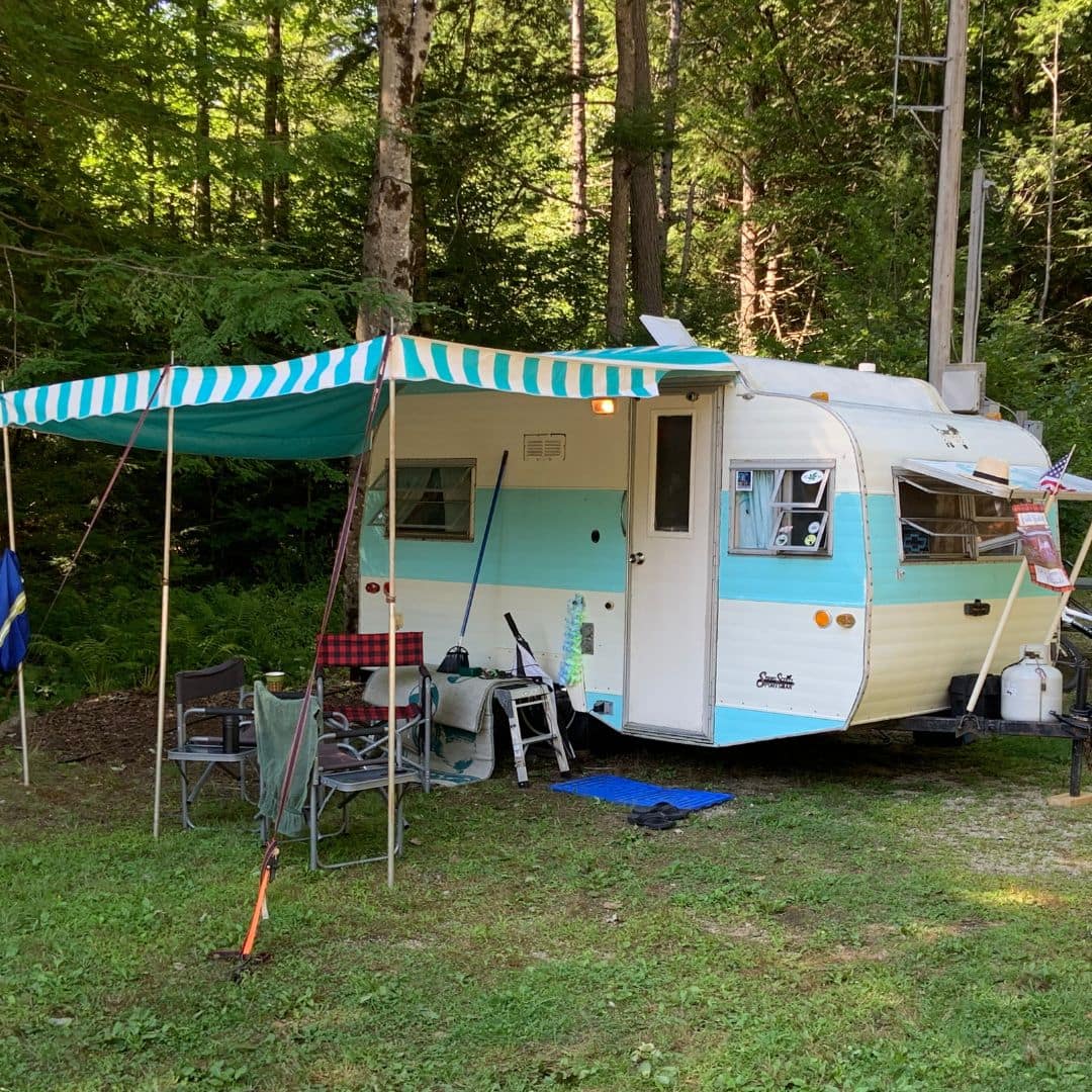 A vintage blue and white camper with a striped canopy is nestled in a grassy area surrounded by trees, under a sky full of galaxies. Outdoor chairs and a small table rest beneath the canopy, evoking a cozy camping scene that invites love and stargazing in this serene wooded setting.