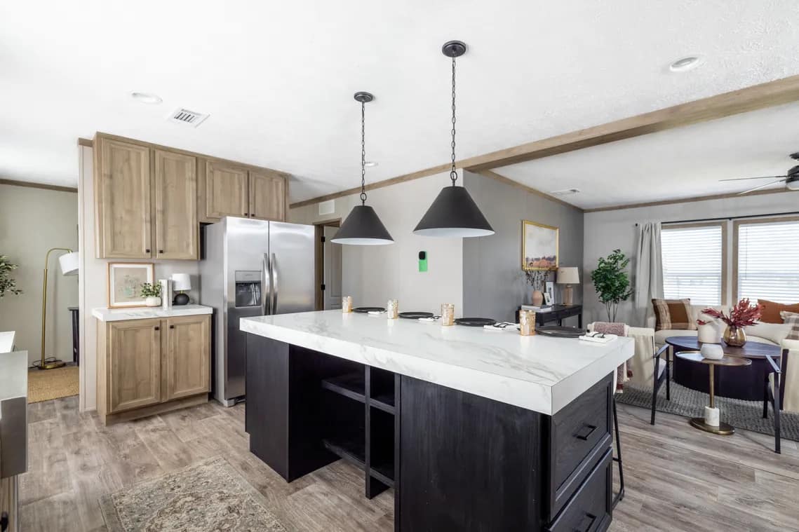Modern kitchen with a large marble island, black pendant lights, stainless steel fridge, light wood cabinets, and an open living area with a sofa and coffee table in the background.