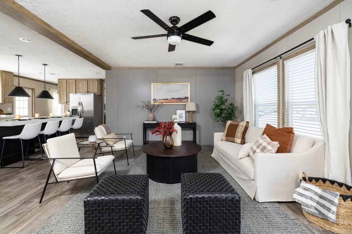 Modern living room with a white sofa, two white chairs, two black ottomans, a round coffee table, neutral decor, large windows with curtains, and an open kitchen in the background.