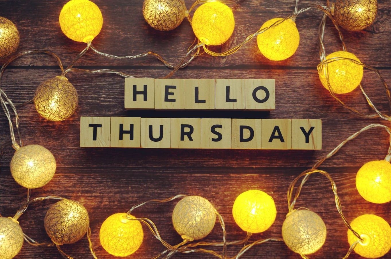Wooden letter blocks spell out "Hello Thursday" on a rustic wooden surface, surrounded by warm yellow string lights shaped like round lanterns—a cozy way to welcome the Third Thursday of the month.