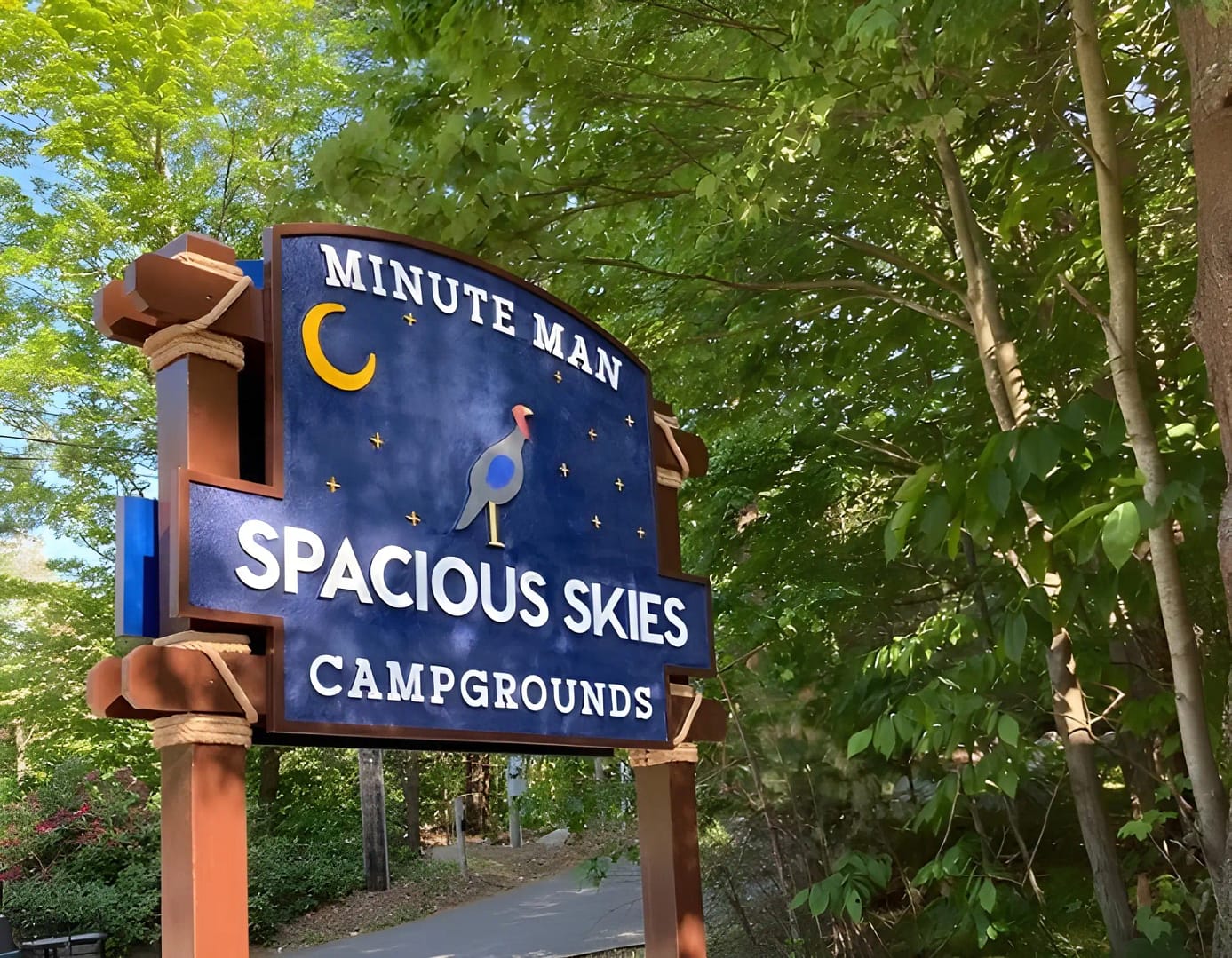 A wooden sign reading "Spacious Skies Minute Man Campgrounds" with a moon and bird illustration, set among green trees, showcases one of the top RV campgrounds near Boston.