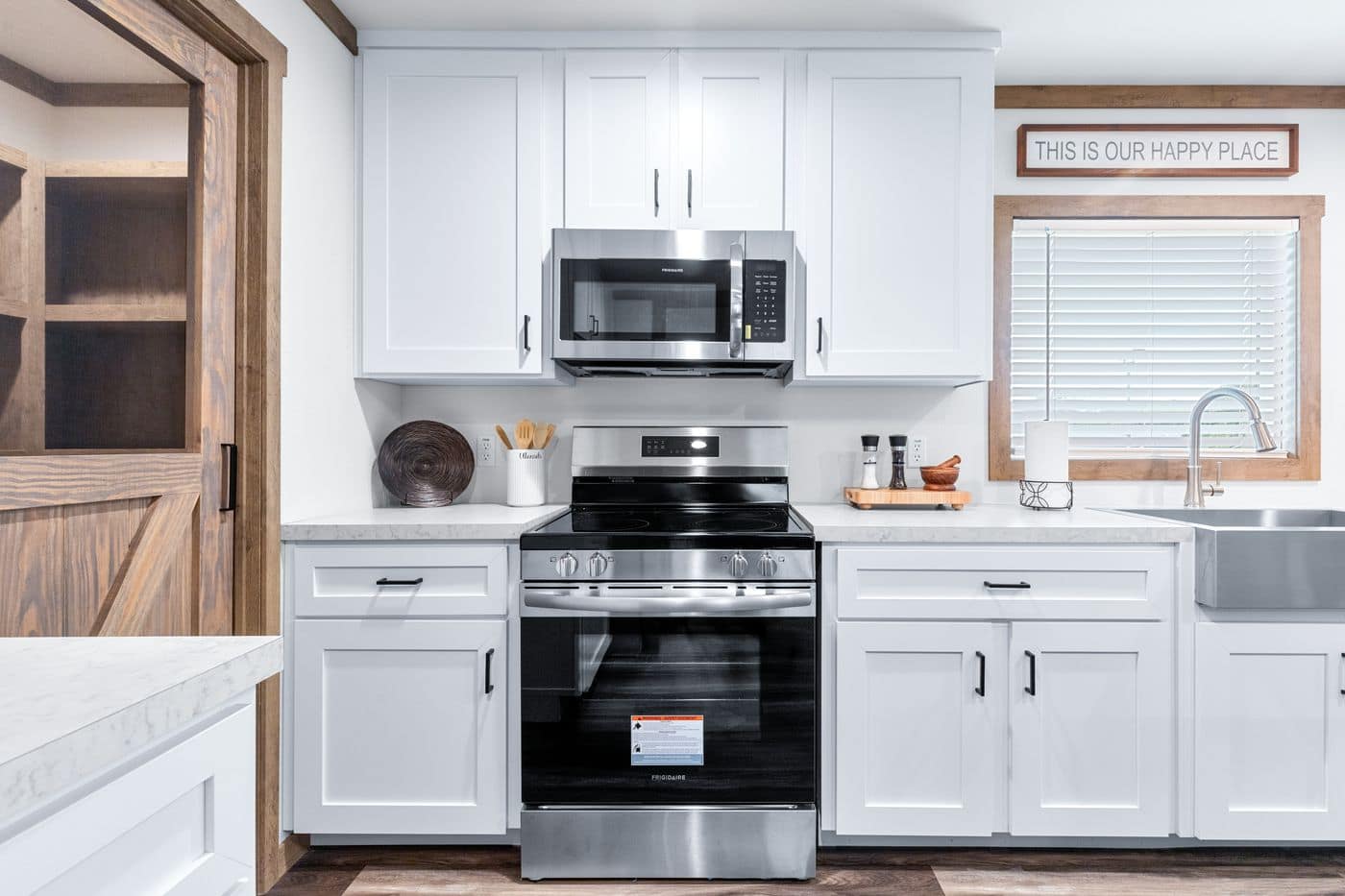 Modern kitchen with white cabinets, stainless steel oven and microwave, farmhouse sink, and a window with blinds. Sign above window reads "This is our happy place.
