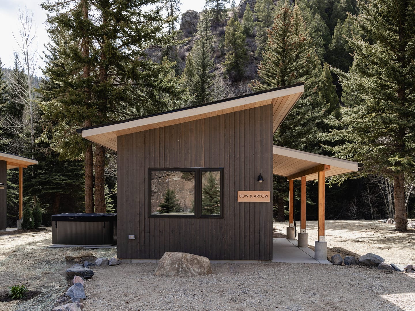A small modern cabin with dark wood siding, large window, and sign reading "BOW & ARROW," set in a forested mountain area with tall evergreen trees and a hot tub to the left.
