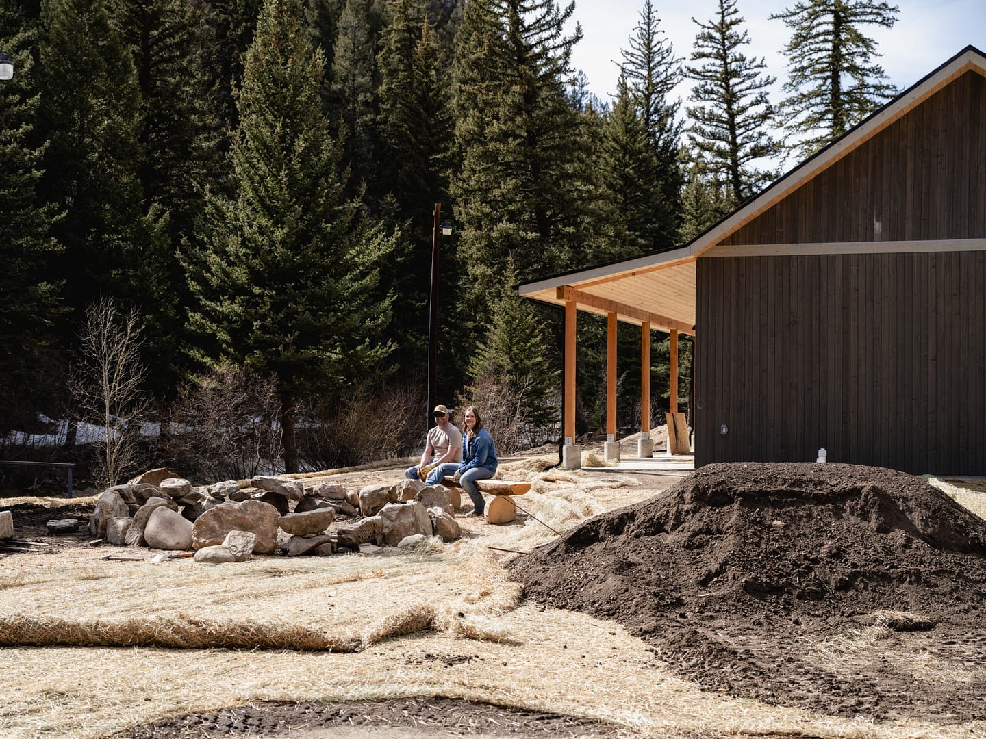 Two people sit on rocks near a modern wooden building surrounded by dirt, straw, and pine trees, with mountains in the background on a sunny day.