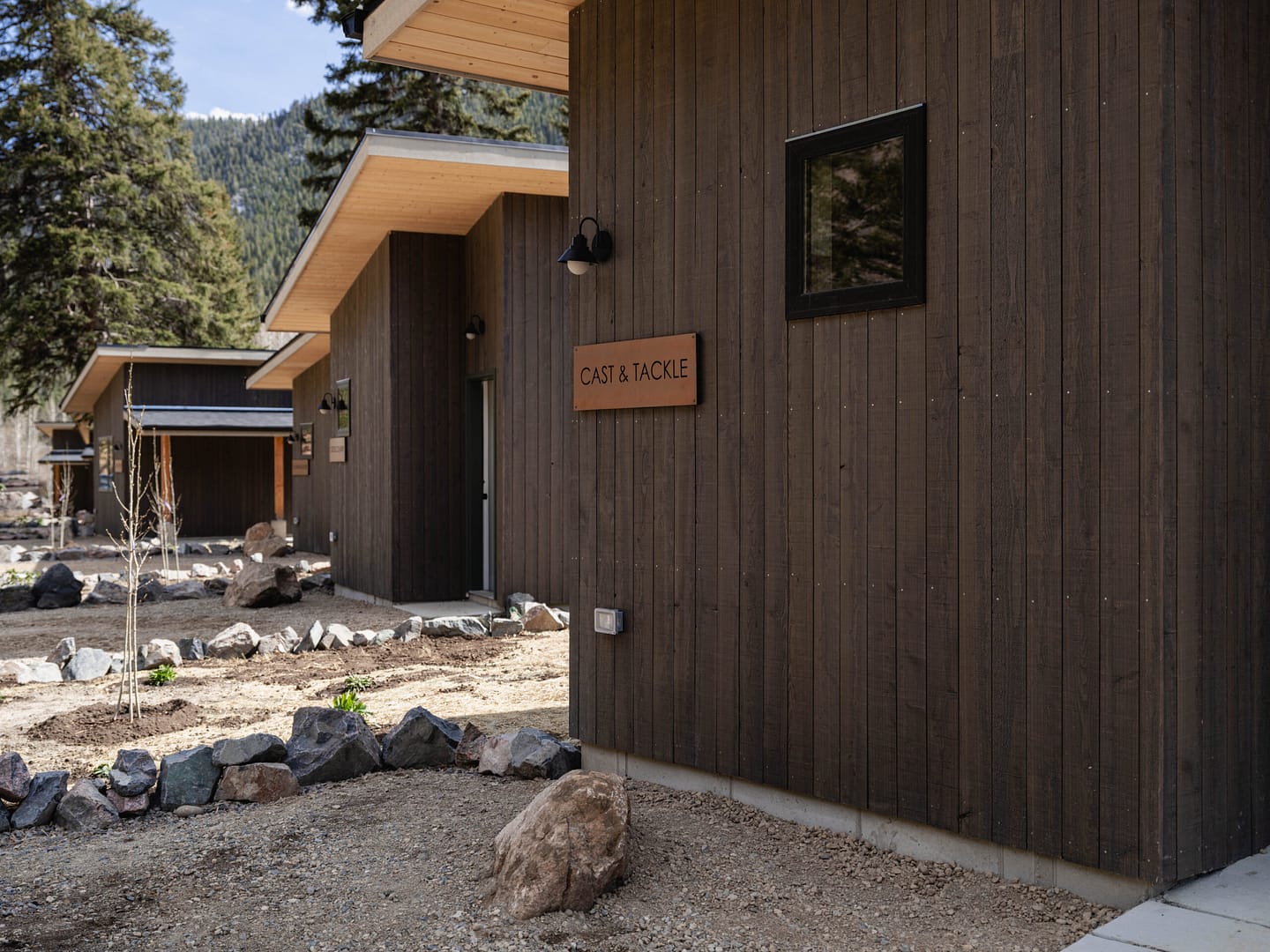 A dark wooden building with a sign reading "Cast & Tackle" is surrounded by rocks, gravel, and trees, with other similar buildings visible in the background under a clear sky.