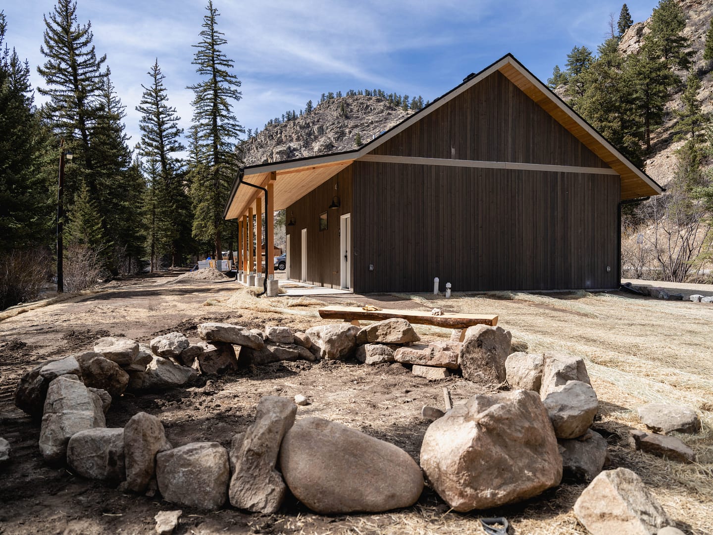 A modern wooden cabin with a sloped roof sits among tall pine trees and mountains. In the foreground, there is a circular fire pit made of large rocks and bare dirt. The sky is clear and sunny.