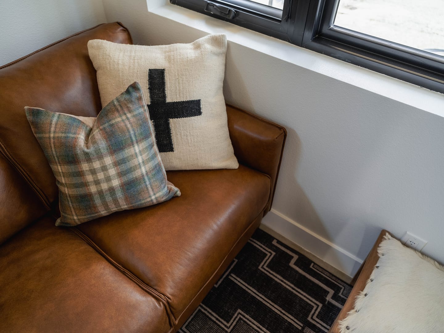 Close-up of a brown leather couch with two decorative pillows—one plaid and one with a large black cross—next to a window and a geometric-patterned rug.
