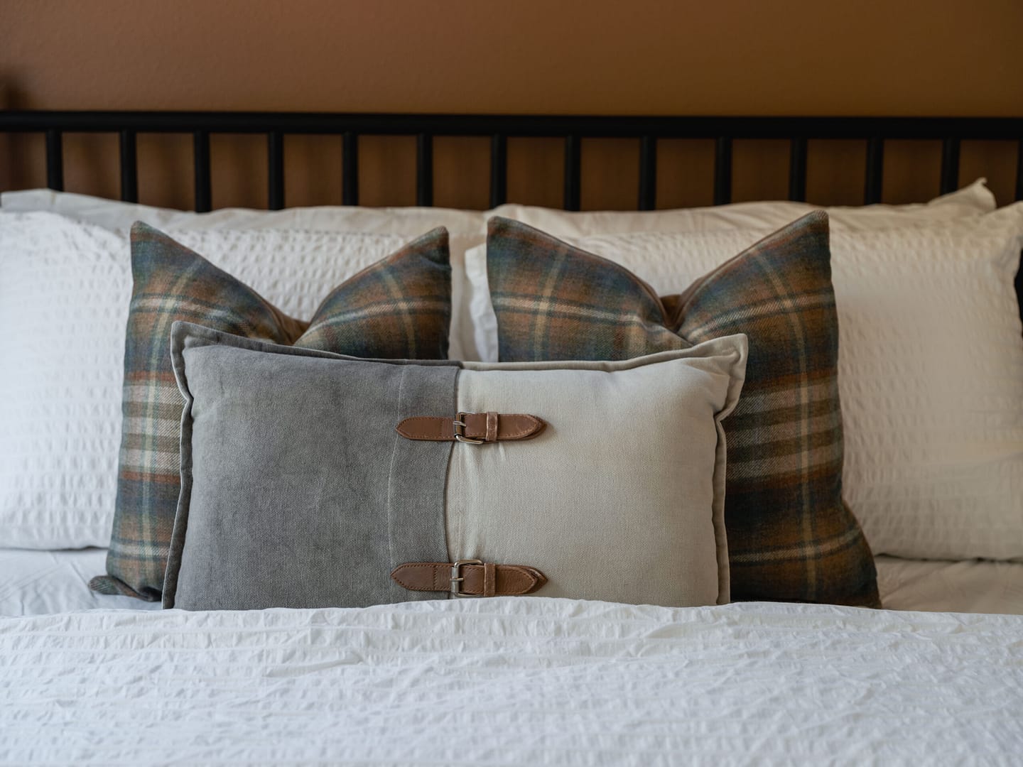 A neatly made bed with white textured bedding, two plaid pillows in shades of green and brown, and a decorative rectangular pillow with gray and beige sections and leather buckle accents in front.