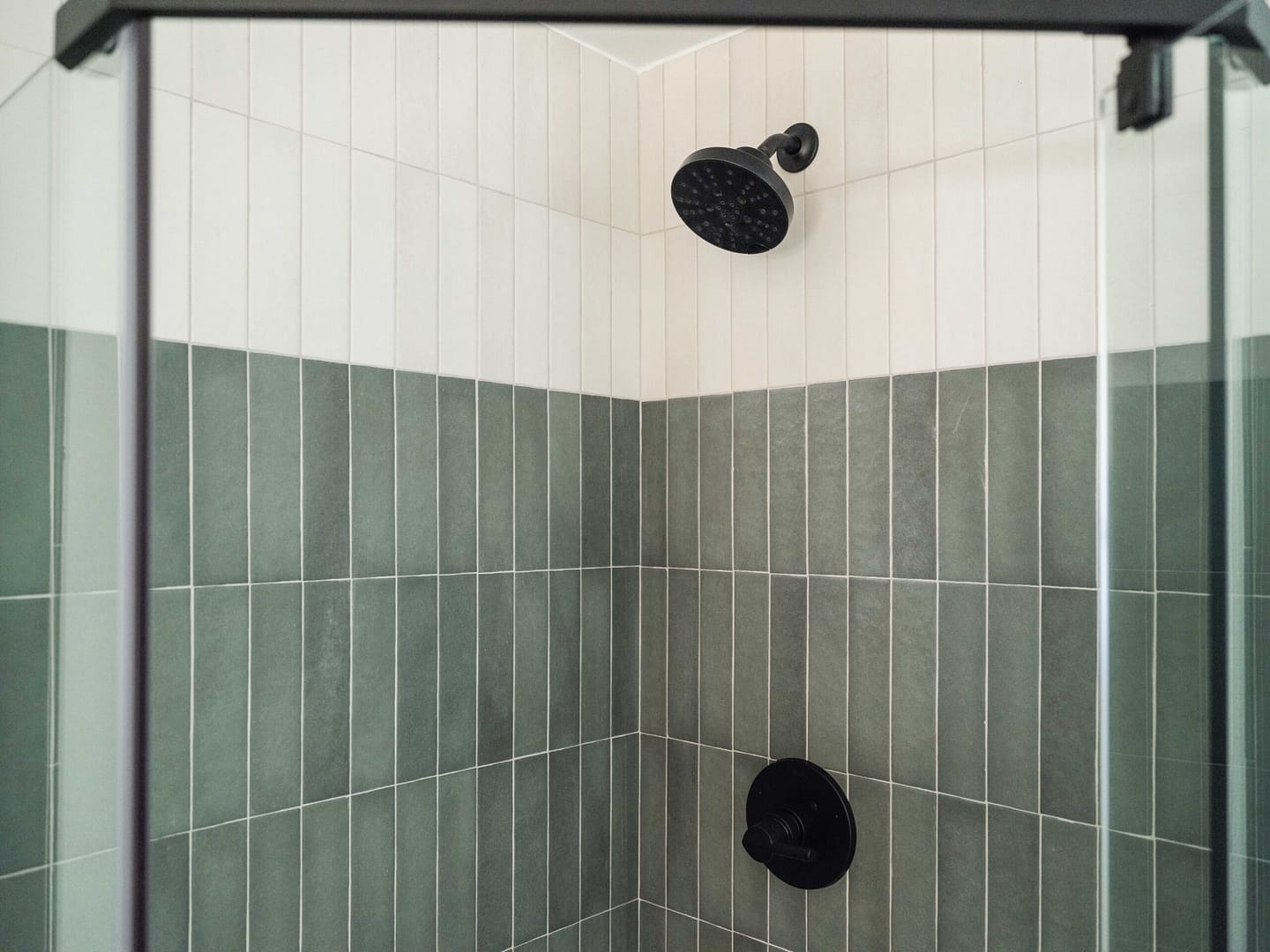 A shower with green and white vertical tile walls, featuring a black showerhead and matching black control knob. The shower is enclosed with glass panels.