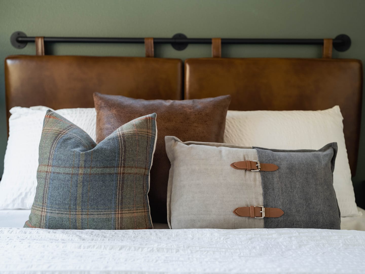 A neatly made bed with white bedding, two brown leather headboards, and three decorative pillows—one plaid, one brown, and one gray with leather buckle accents—arranged in front.