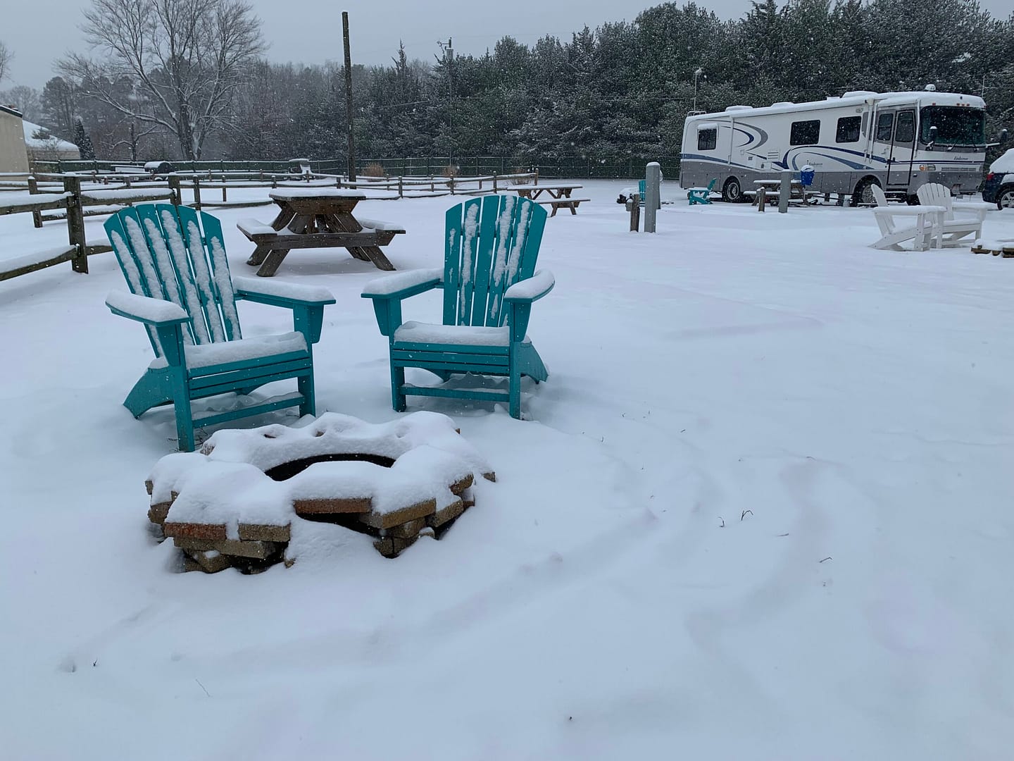 Two turquoise Adirondack chairs sit in the snow around a fire pit, inviting you to stay cozy. A picnic table nearby is dusted with snow. In the background, an RV rests at Spacious Skies Campgrounds, and trees line the horizon under a gray winter sky—a perfect winter camping scene.