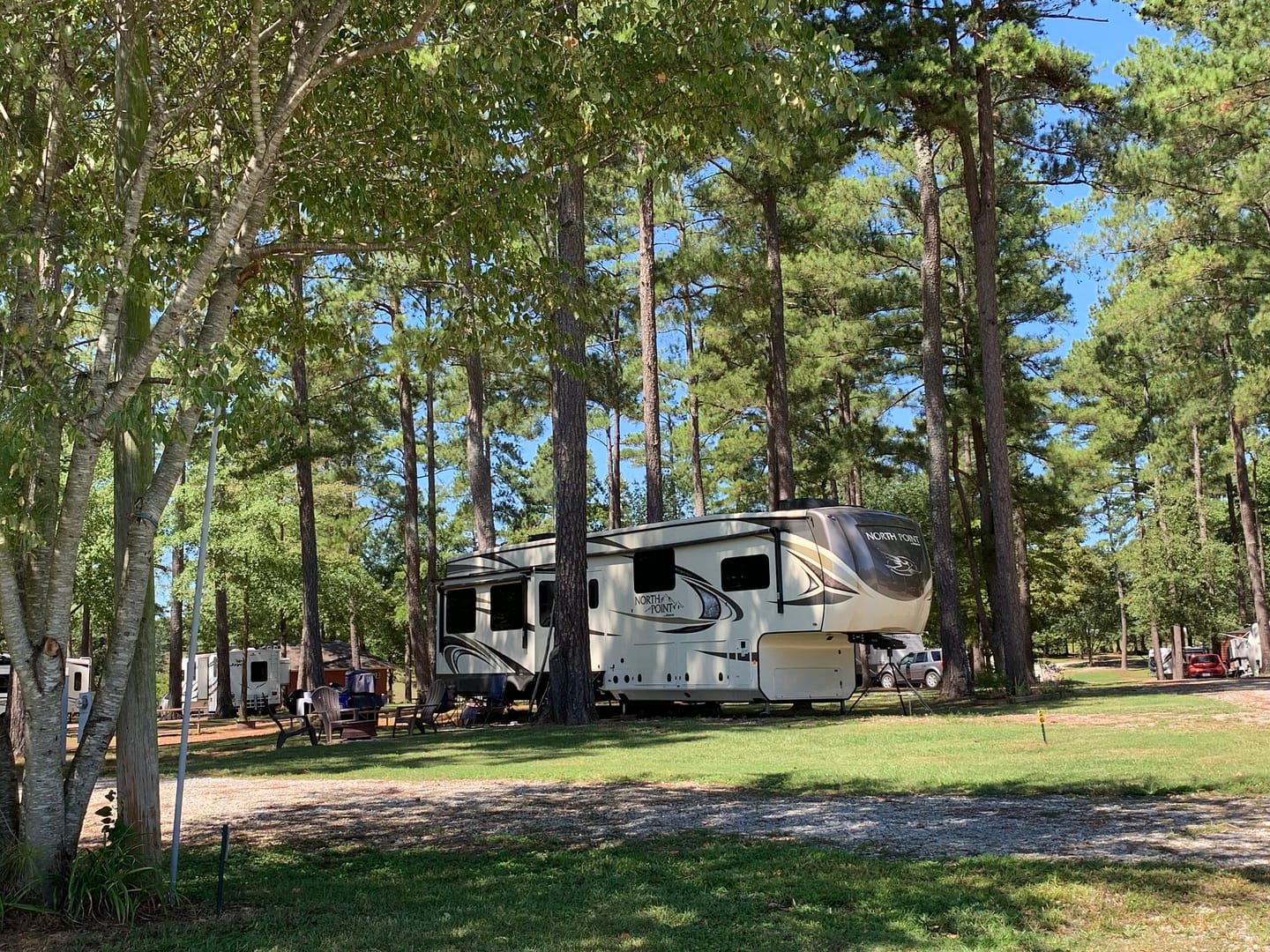A large RV is parked among tall pine trees at a sunny campground in Spartanburg SC, with picnic tables and other vehicles visible in the background—ideal for long-term camping.