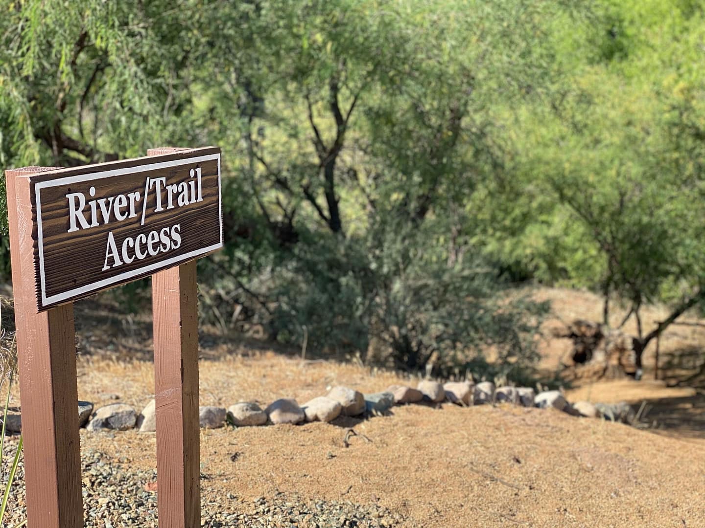 A wooden sign reads "River Trail Access" beside a dirt path surrounded by trees and rocks in a natural outdoor setting.