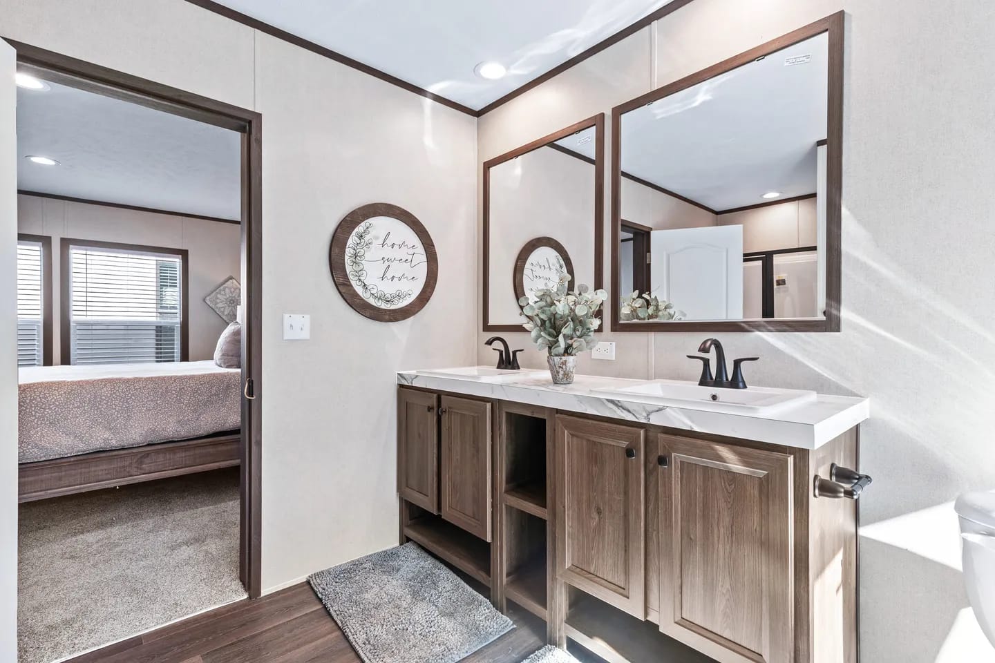 Modern bathroom with double sinks, two large mirrors, wood cabinets, decorative wall art, and a view into a bedroom with a bed and window blinds.