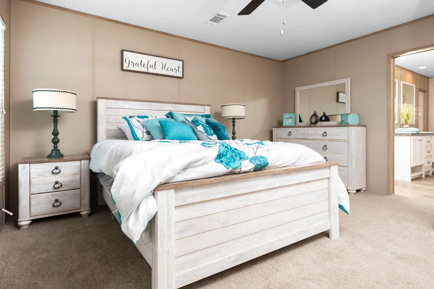 A bedroom with a wooden bed frame, teal and white bedding, two nightstands with lamps, a dresser with a mirror, and beige carpeted floor.