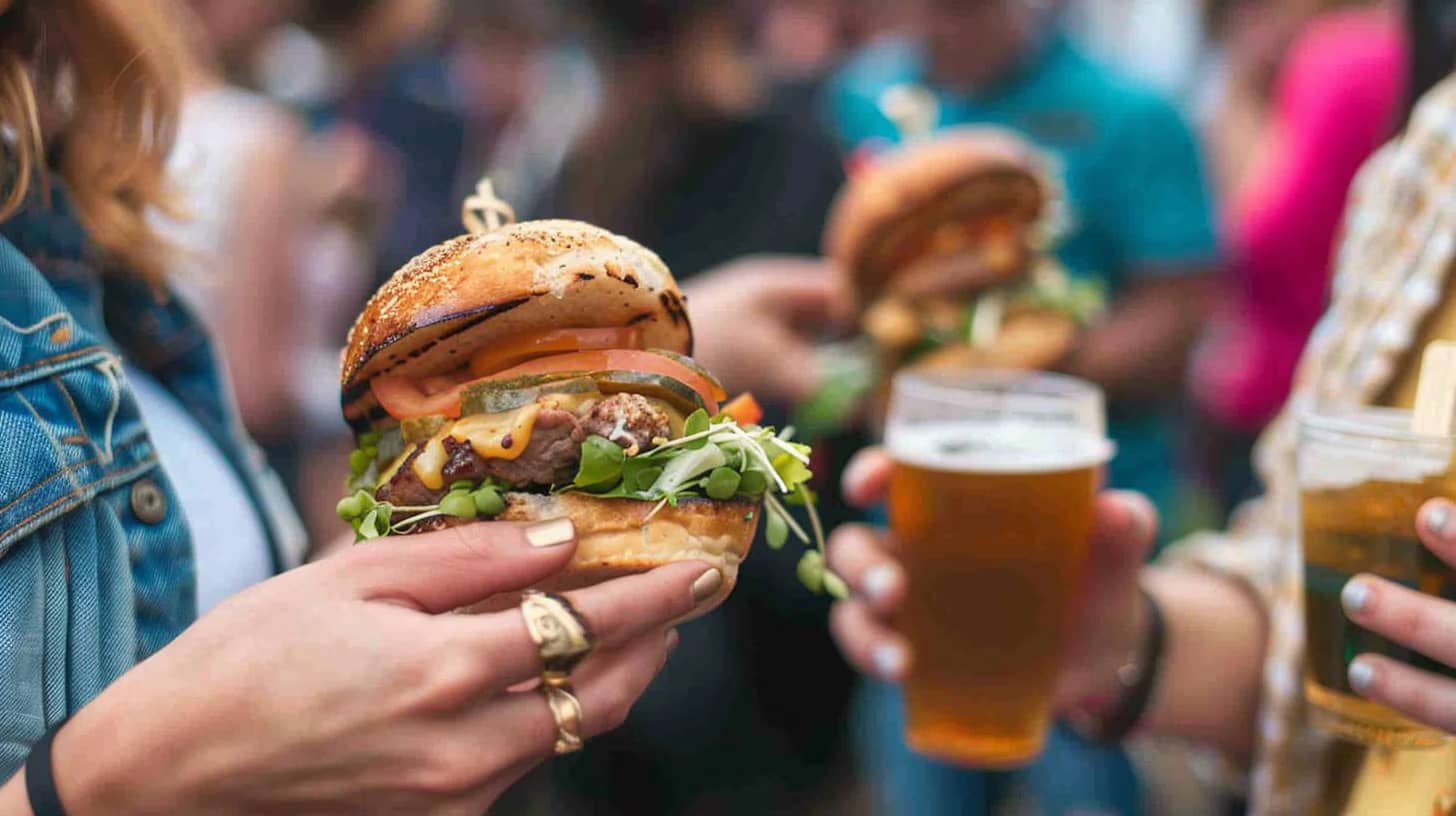 A person holds a large, gourmet burger with lettuce, cheese, and pickles, while another person holds a glass of beer. The background shows a crowd of people, suggesting a casual outdoor gathering.