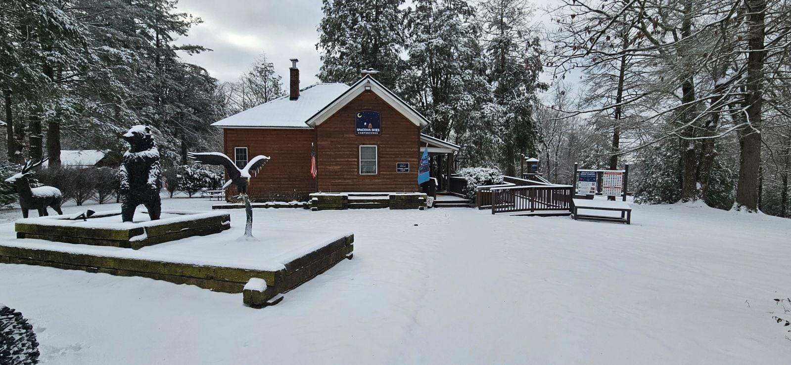 A snowy scene with a wooden cabin, flanked by large trees. In front, statues of a bear and a bird stand on a snow-covered platform. Signs are visible near the entrance, and the ground is blanketed in fresh snow.
