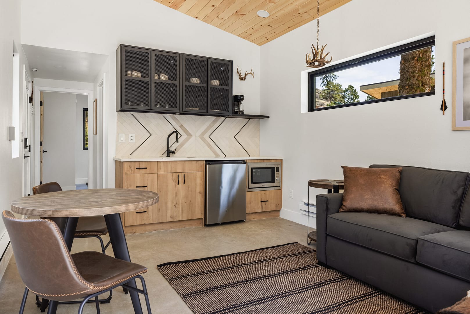 A modern studio with a small kitchen featuring light wood cabinets, a mini fridge, a round dining table with two chairs, a black sofa, and a rug. The room has a wood ceiling and a window letting in natural light.