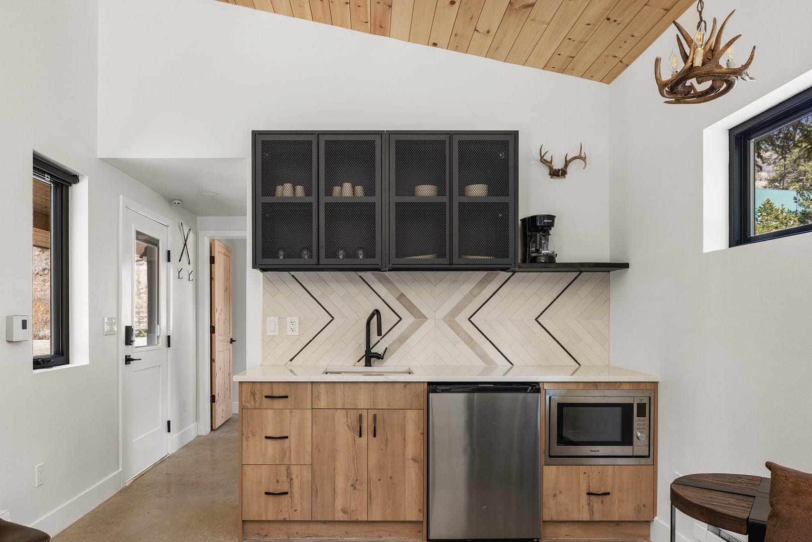 Modern kitchenette with wood cabinets, black fixtures, a geometric tile backsplash, glass-front upper cabinets, microwave, small refrigerator, antler decor, and natural light from windows.