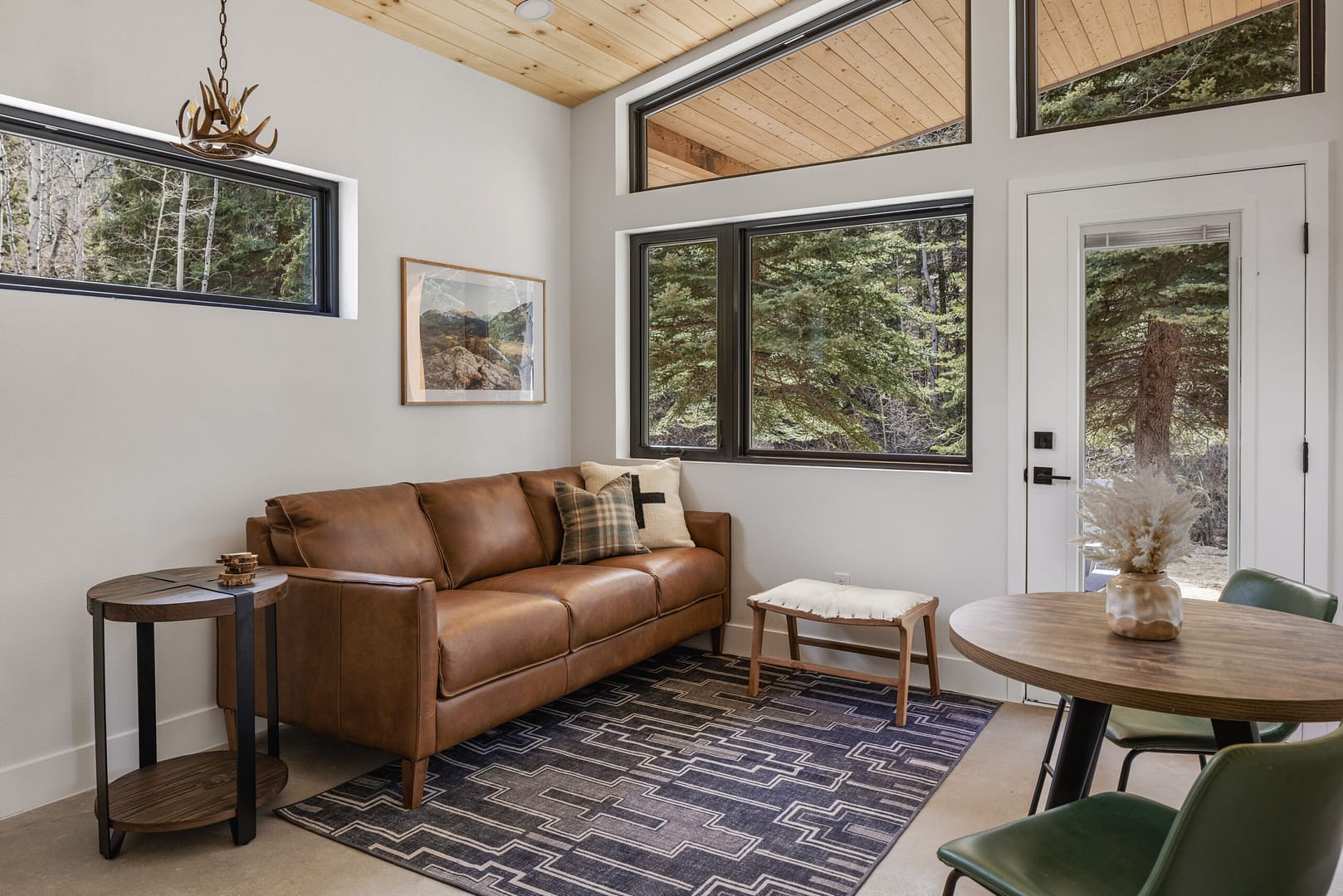 Cozy living room with a brown leather sofa, patterned rug, round side table, small bench, and a round dining table with green chairs. Large windows offer views of trees and let in natural light.