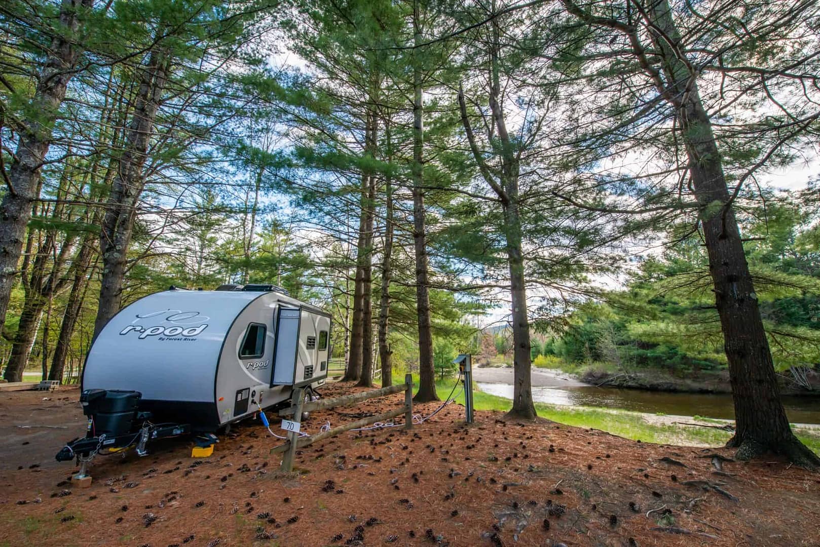 An RV parked in the woods near a river, surrounded by blooming flowers.