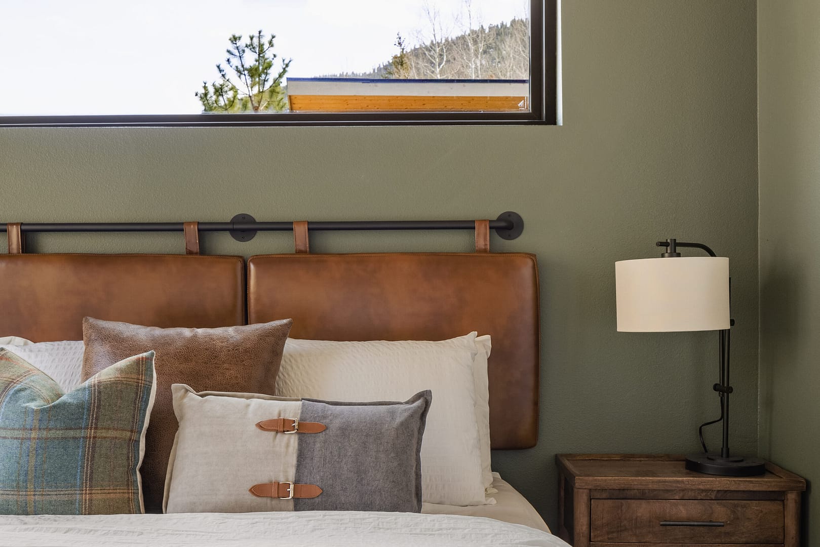 A cozy bedroom with a brown leather headboard, decorative pillows in neutral and plaid patterns, a wooden nightstand, a black lamp, and a window showing a view of trees and mountains.