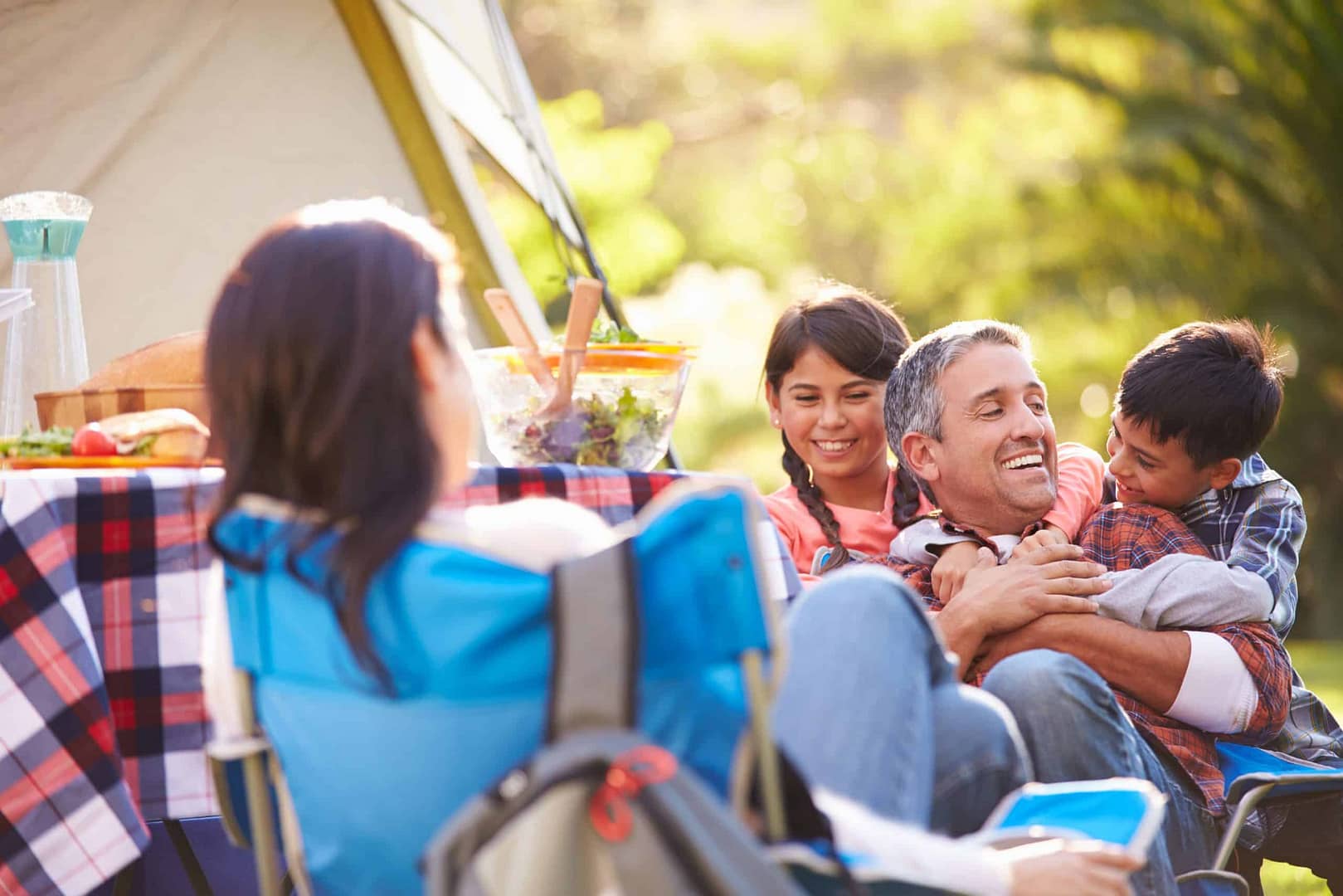 young kids hugging dad on camping trip near tent and picnic table