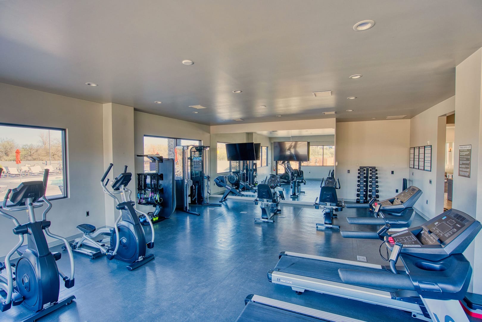 Modern gym with treadmills, stationary bikes, weight machines, free weights, and large mirrors; bright with natural light from windows.