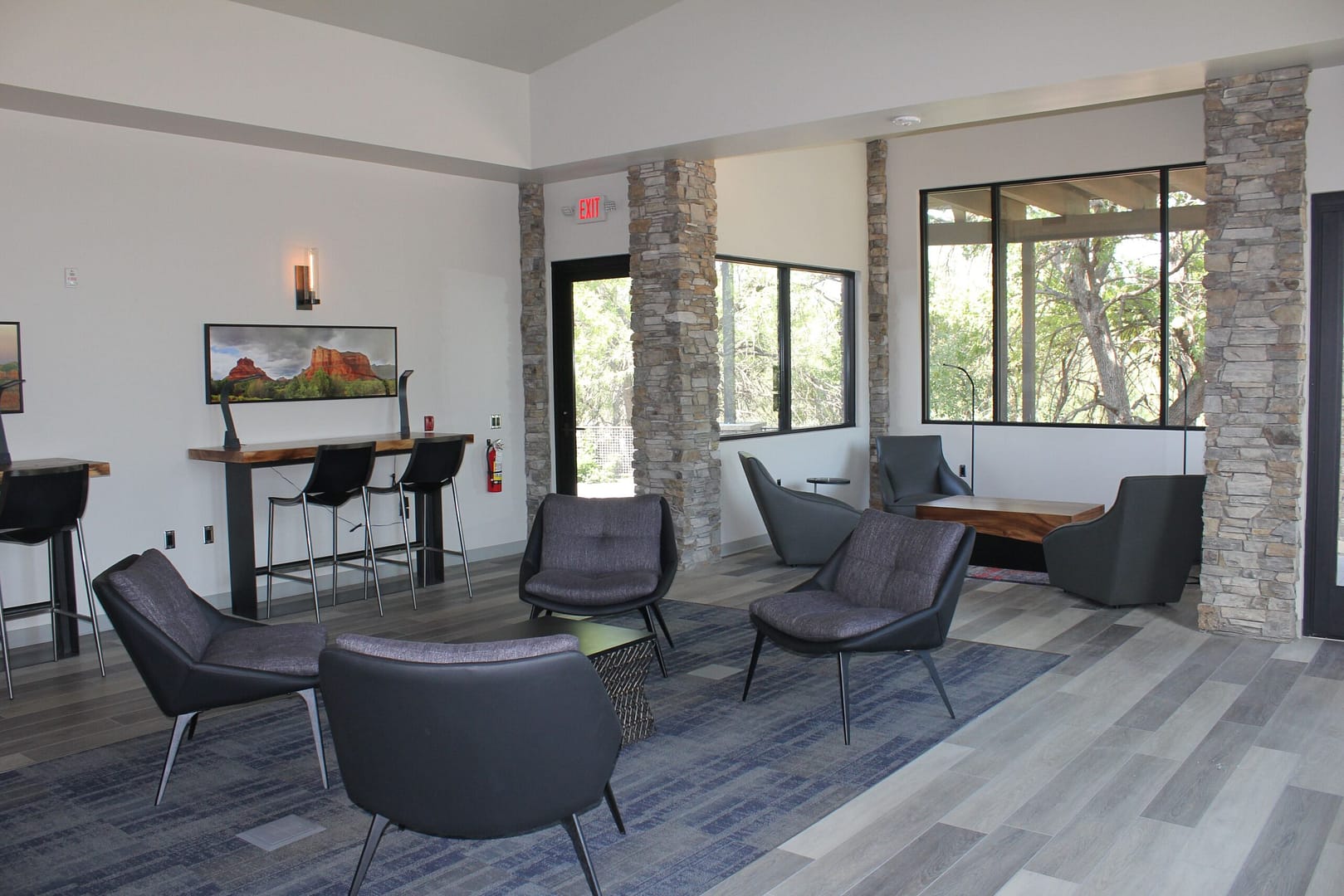 Modern lounge area with gray chairs, a low table, bar seating, large windows, stone pillars, and wood-style flooring.