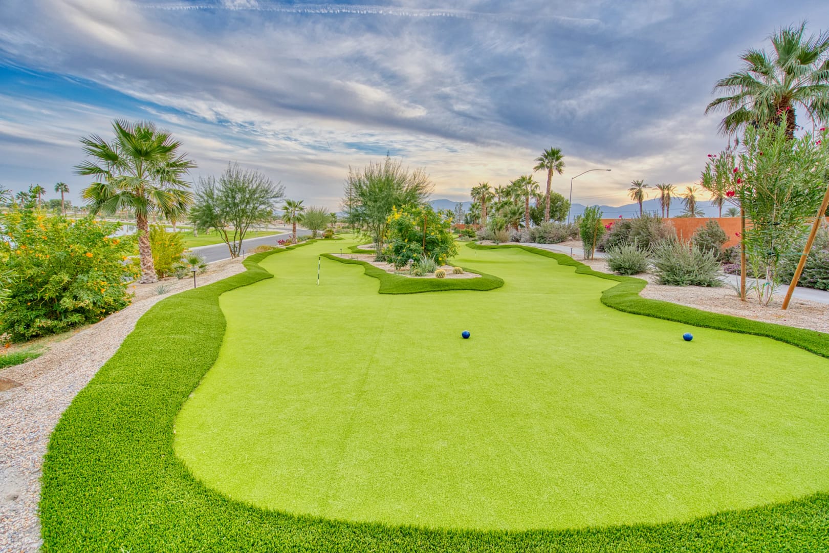 A landscaped mini-golf course with putting greens, surrounding plants, palm trees, and a partly cloudy sky.
