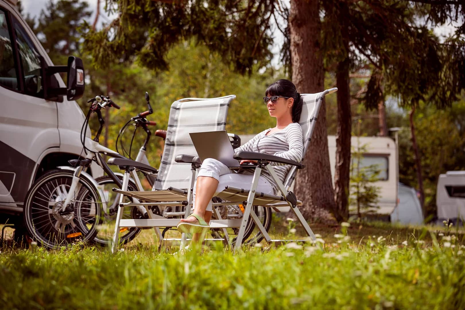 A woman sitting in a lawn chair with a laptop in front of an RV enjoying the great outdoors.