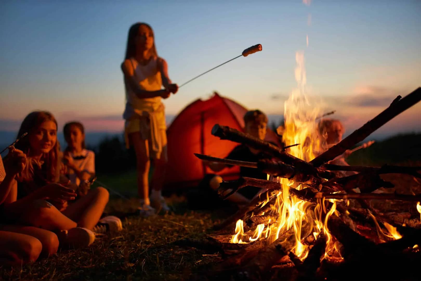 A group of people sit around a campfire at dusk, with one person standing and roasting marshmallows on a stick. An orange tent is set up in the background, creating the perfect setting for an impromptu auto draft of adventure stories.